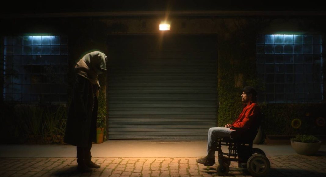 Pessoa encapuzada e homem em cadeira de rodas, à noite.