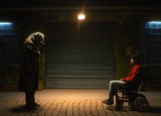 Pessoa encapuzada e homem em cadeira de rodas, à noite.