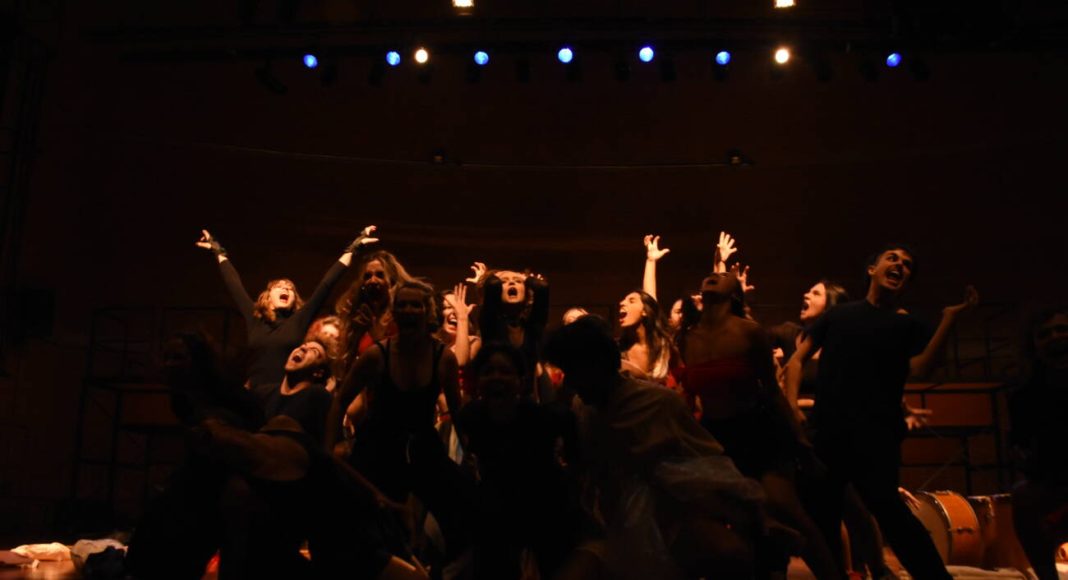 Grupo teatral em cena animada no palco iluminado.