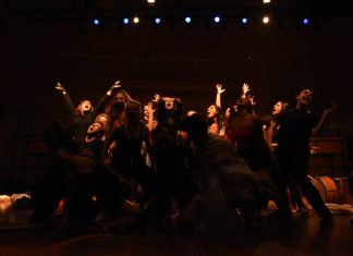 Grupo teatral em cena animada no palco iluminado.