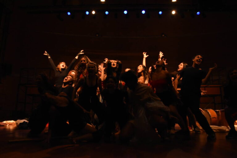 Grupo teatral em cena animada no palco iluminado.