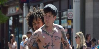 Homem tatuado na rua com pessoas ao fundo.