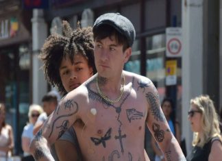 Homem tatuado na rua com pessoas ao fundo.