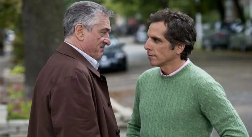 Two men stand face-to-face on a street, one in a brown coat and the other in a green knit sweater, engaged in a serious conversation.