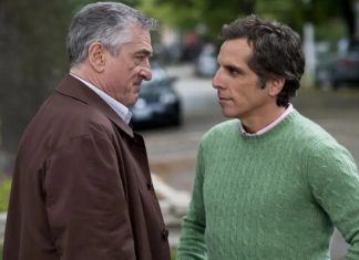 Two men stand face-to-face on a street, one in a brown coat and the other in a green knit sweater, engaged in a serious conversation.