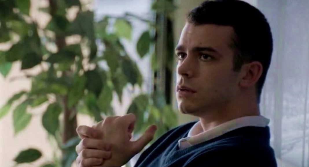 A man sits indoors by a window with green plants, hands clasped in front, looking thoughtful or pensive.