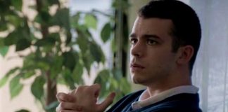 A man sits indoors by a window with green plants, hands clasped in front, looking thoughtful or pensive.