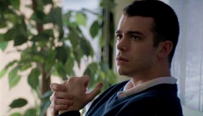 A man sits indoors by a window with green plants, hands clasped in front, looking thoughtful or pensive.