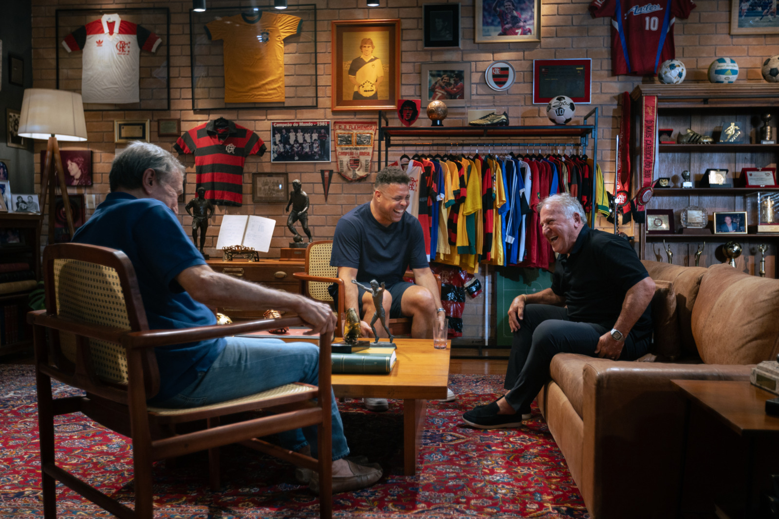 Three men sit and laugh around a wooden coffee table in a sports-filled living room with vintage jerseys on display behind them.