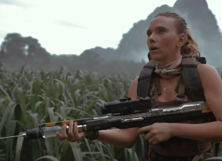 Woman in tactical vest and scarf holding a scoped sniper rifle in a dense grassy field, looking alert.