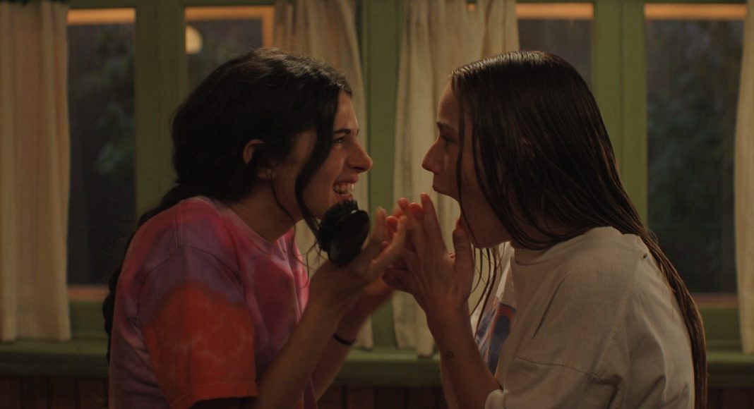 Sterling Point_ Ella Rubin-Amélie Elisabeth Hoeferle Two young women lean in close, whispering and smiling at each other in a cozy room with curtains in the background.