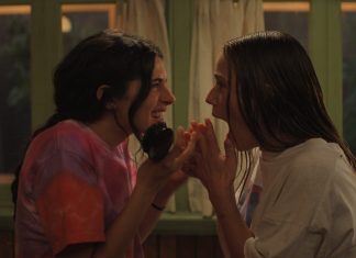 Two young women lean in close, whispering and smiling at each other in a cozy room with curtains in the background.