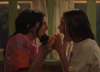 Segredos e romances nas cenas inéditas de ‘Sterling Point’, novo DRAMA do Prime Video! Two young women lean in close, whispering and smiling at each other in a cozy room with curtains in the background.