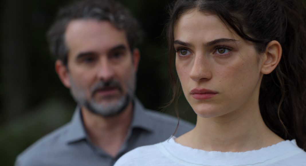 Close-up of a young woman with a serious expression; a blurred man stands in the background outdoors.
