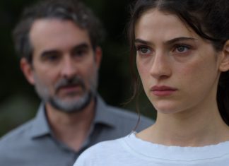 Close-up of a young woman with a serious expression; a blurred man stands in the background outdoors.