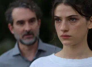 Segredos e romances no clipe inédito de ‘Sterling Point’, novo DRAMA do Prime Video! Close-up of a young woman with a serious expression; a blurred man stands in the background outdoors.