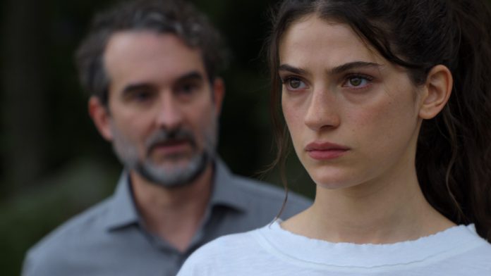 Close-up of a young woman with a serious expression; a blurred man stands in the background outdoors.