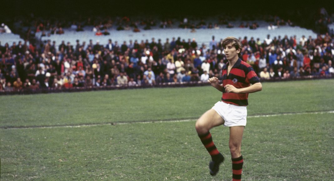 Rugby player in red and black striped jersey and white shorts runs on a grassy field with a crowded stadium in the background.]}