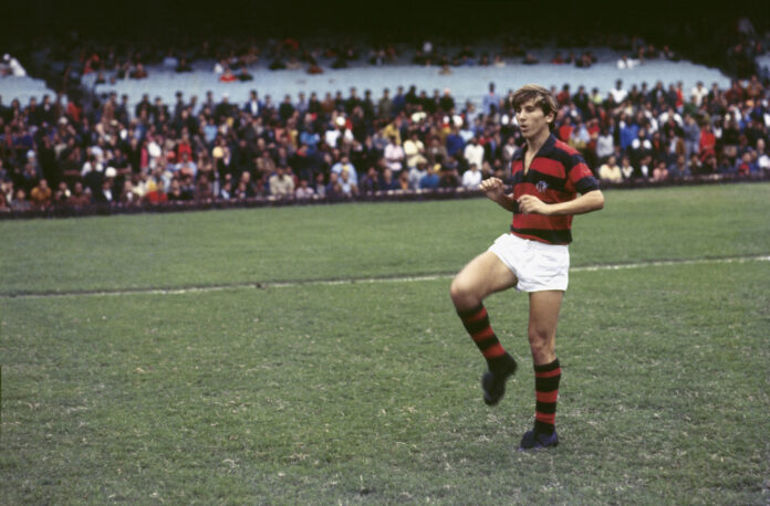 Rugby player in red and black striped jersey and white shorts runs on a grassy field with a crowded stadium in the background.]}