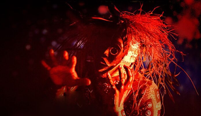 A masked, horror-style figure with straw-like hair reaching toward the camera in red lighting.