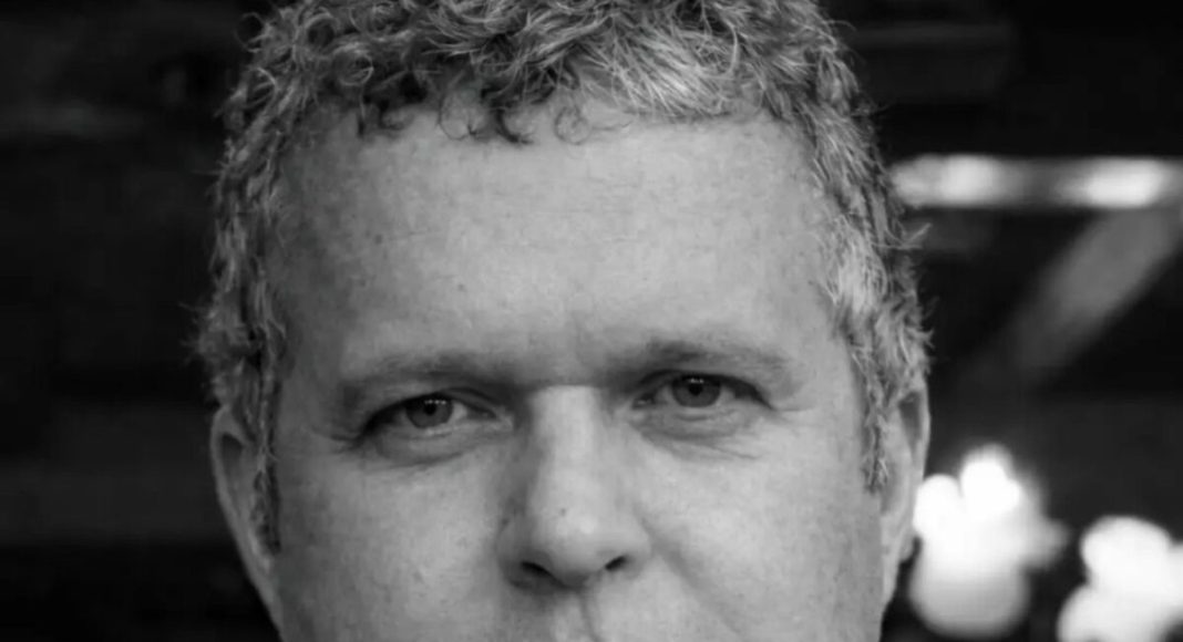 Black-and-white portrait of a man with short, curly gray hair wearing a dark T-shirt, looking at the camera.