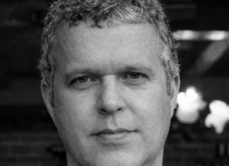 Black-and-white portrait of a man with short, curly gray hair wearing a dark T-shirt, looking at the camera.