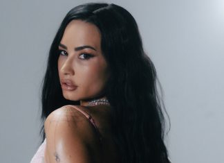 Woman with long black hair looks over her shoulder, wearing a pink top and a beaded necklace against a gray background.
