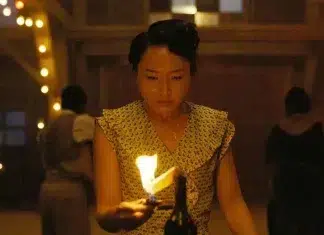 Atriz de ‘Pecadores’ se junta ao elenco da 3ª temporada de ‘The Last of Us’ Woman in a patterned dress holds a lit candle in a warm, dim room with string lights in the background.