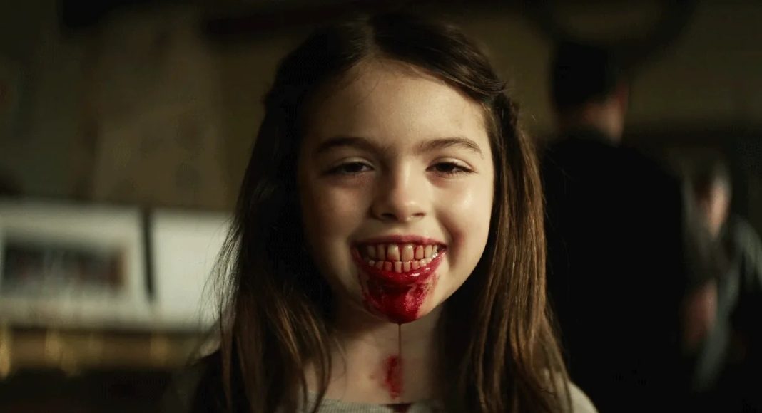 Young girl smiling with blood on her lips and chin in a dimly lit room, suggesting a horror or Halloween scene.