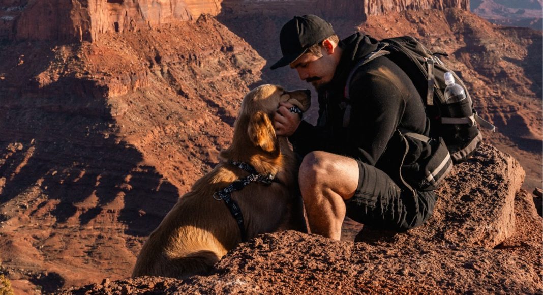 Man in a black cap kneels on a red rock ledge, gently holding a golden retriever's face for a kiss during a canyon hike, backpack nearby.