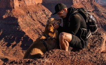 Man in a black cap kneels on a red rock ledge, gently holding a golden retriever's face for a kiss during a canyon hike, backpack nearby.