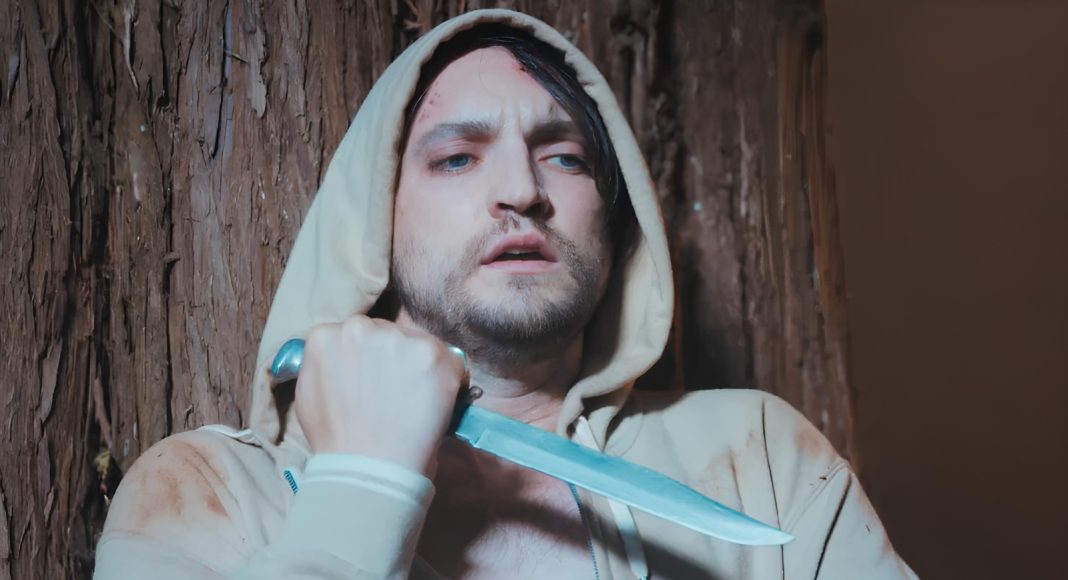 Man in a beige hoodie holding a large knife against his neck, looking distressed beside a tree trunk.