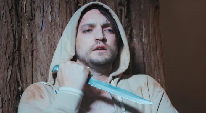 Man in a beige hoodie holding a large knife against his neck, looking distressed beside a tree trunk.
