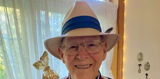 Smiling elderly man in a striped red, white, and blue shirt wearing a straw hat with a blue band; a butterfly rests near his shoulder.