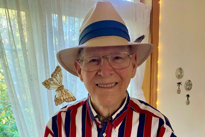 Smiling elderly man in a striped red, white, and blue shirt wearing a straw hat with a blue band; a butterfly rests near his shoulder.