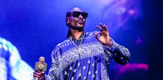 Male performer on stage wearing a blue paisley-pattern shirt and sunglasses, holding a glittering microphone.