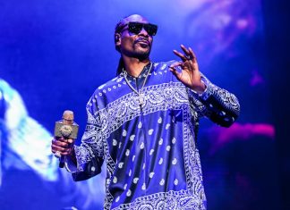 ‘Snoop’: Cinebiografia de Snoop Dogg ganha data de estreia nos cinemas dos EUA; Confira! Male performer on stage wearing a blue paisley-pattern shirt and sunglasses, holding a glittering microphone.
