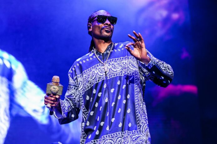Male performer on stage wearing a blue paisley-pattern shirt and sunglasses, holding a glittering microphone.
