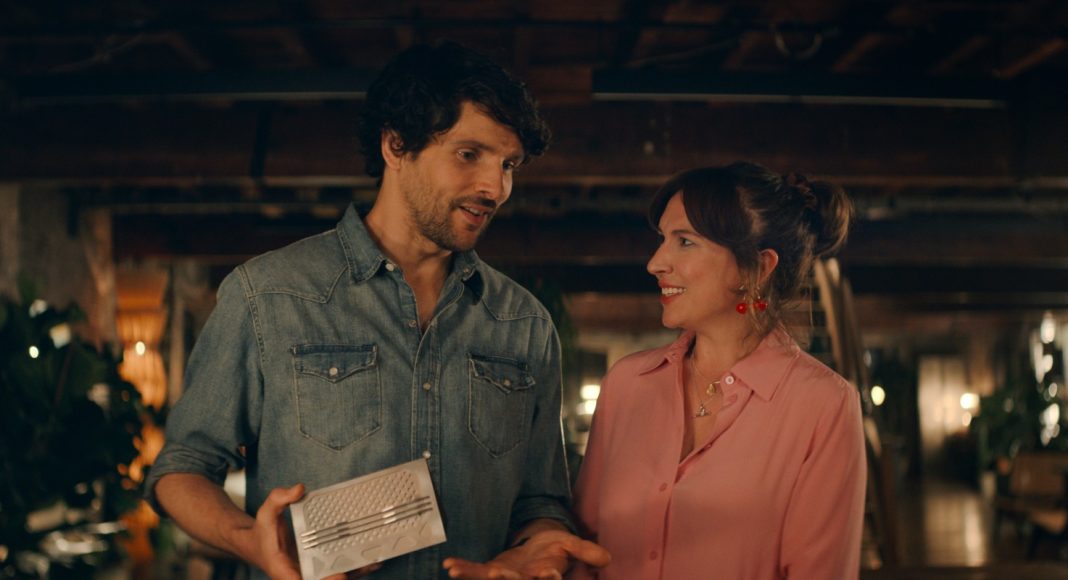 Two adults in a dim, cozy space smile at each other as the man shows a silver rectangular gadget to the woman.