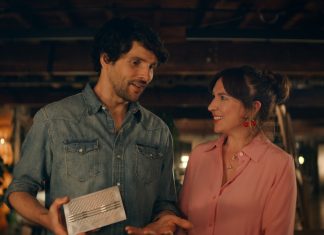 Two adults in a dim, cozy space smile at each other as the man shows a silver rectangular gadget to the woman.
