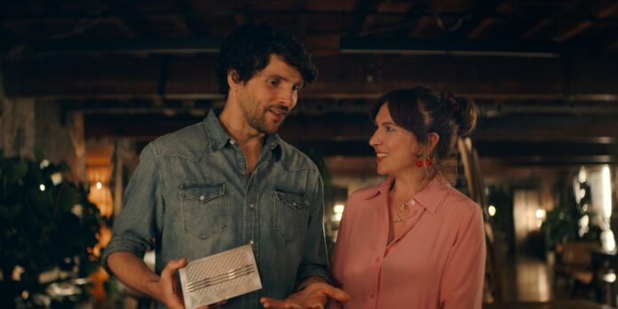 Two adults in a dim, cozy space smile at each other as the man shows a silver rectangular gadget to the woman.