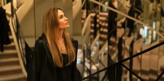 Woman with long blonde hair wearing a black coat, standing near a railing inside a modern building with stairs in the background, looking to the left.