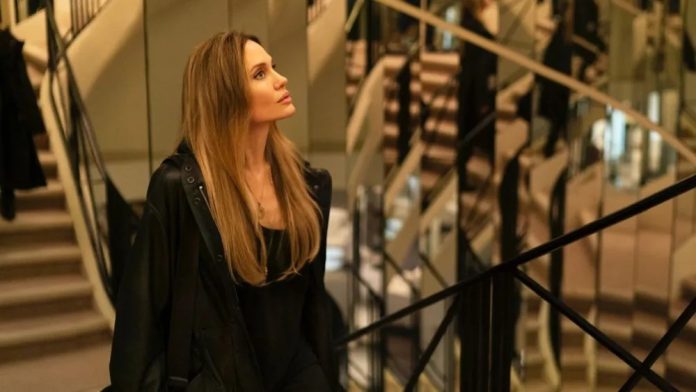 Woman with long blonde hair wearing a black coat, standing near a railing inside a modern building with stairs in the background, looking to the left.