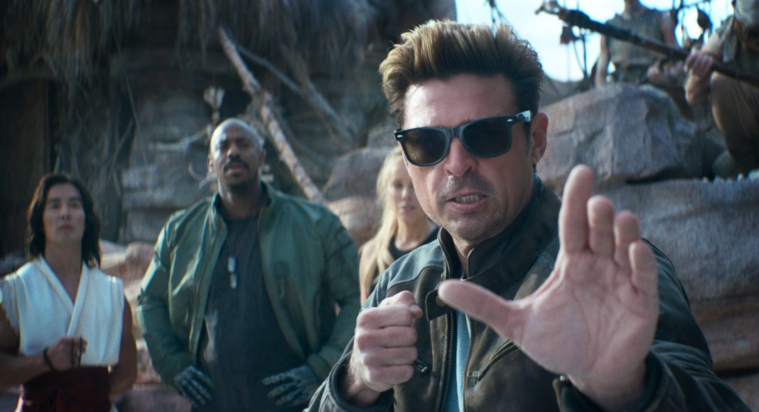 Man in sunglasses and a brown leather jacket extends his hand toward the camera as if stopping you, while three people stand behind him in a rocky outdoor setting.