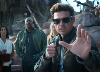 Man in sunglasses and a brown leather jacket extends his hand toward the camera as if stopping you, while three people stand behind him in a rocky outdoor setting.