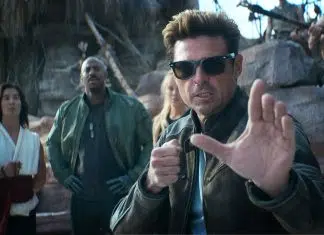 Man in sunglasses and a brown leather jacket extends his hand toward the camera as if stopping you, while three people stand behind him in a rocky outdoor setting.