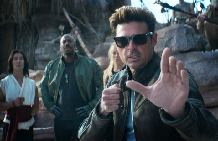 Man in sunglasses and a brown leather jacket extends his hand toward the camera as if stopping you, while three people stand behind him in a rocky outdoor setting.