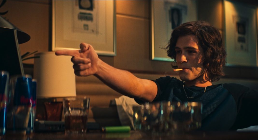 Man with shoulder-length hair sits at a dimly lit bar, cigarette in mouth, pointing off-screen with his right hand and raising his left hand in the air.