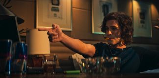 Man with shoulder-length hair sits at a dimly lit bar, cigarette in mouth, pointing off-screen with his right hand and raising his left hand in the air.