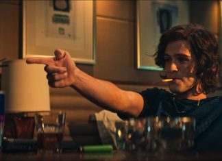 Man with shoulder-length hair sits at a dimly lit bar, cigarette in mouth, pointing off-screen with his right hand and raising his left hand in the air.
